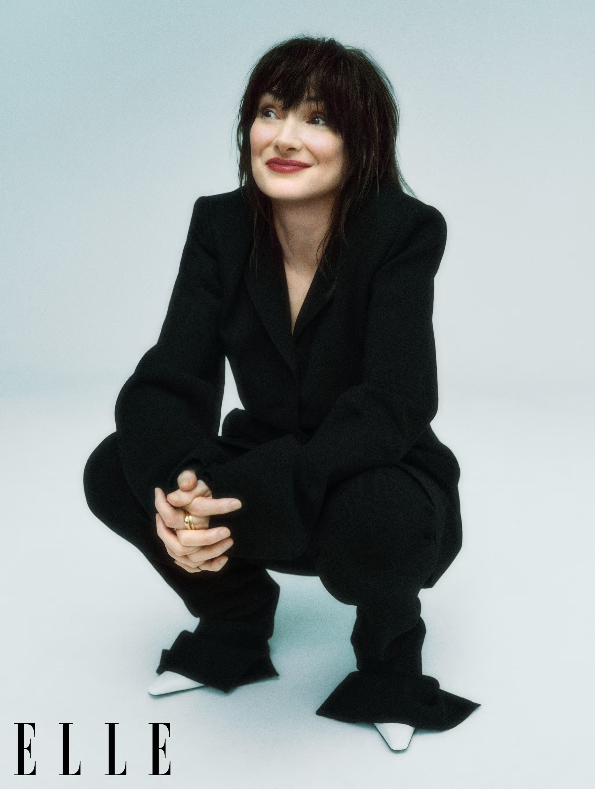 Woman in a black pantsuit with white shoes crouches in a photo studio against a light blue backdrop, gazing off camera with a slight smile.