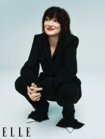 Woman in a black pantsuit with white shoes crouches in a photo studio against a light blue backdrop, gazing off camera with a slight smile.