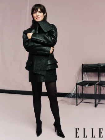 Winona Ryder in a black textured jacket and skirt with tights standing in a studio for Elle UK