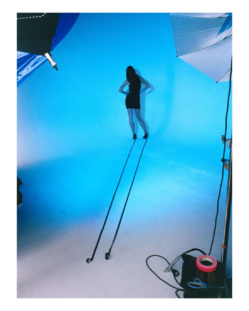 Model Julia Banas in a black dress standing against a vibrant blue studio backdrop with long dramatic shadows cast forward.