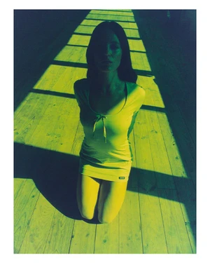 Model kneeling on a wooden floor with stark linear shadows cast across the scene in high-contrast blue and yellow tones.