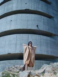 Model standing on rocky terrain wearing a structured beige coat against a large curved concrete structure.