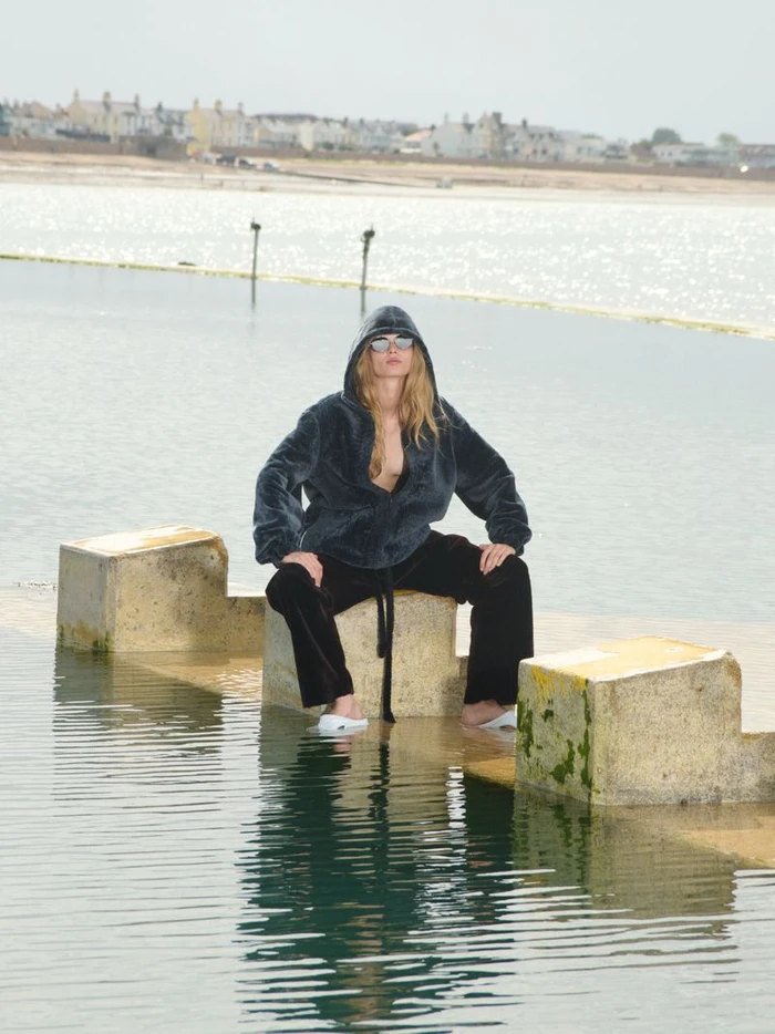 Model wearing a dark hooded fleece and black trousers seated on a concrete structure in water for Self Service 65.