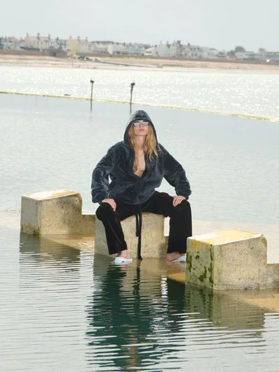 Model wearing a dark hooded fleece and black trousers seated on a concrete structure in water for Self Service 65.