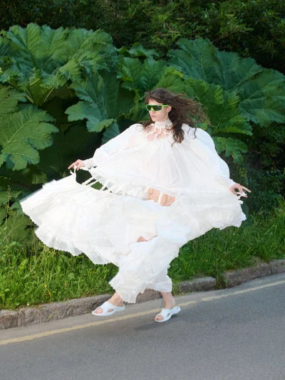 Model in voluminous white dress and green sunglasses posing outdoors before large green leaves for Self Service