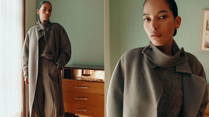 Model in taupe wool coat and matching trousers posing in a domestic interior with green walls and wooden dresser