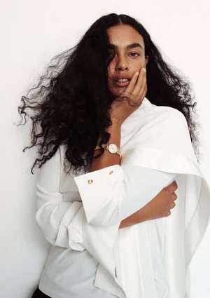 Model with long dark curly hair wearing a white top and gold watch against a plain white background for Temps limite.