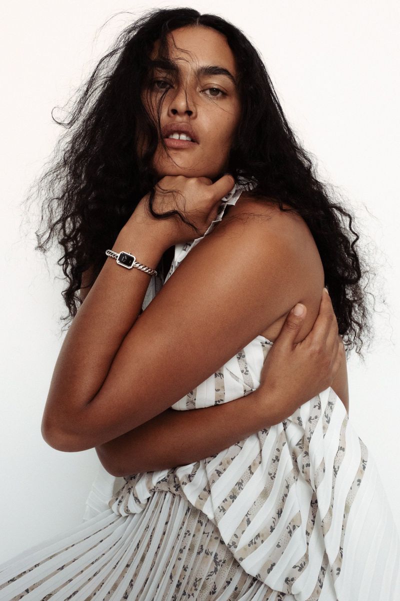 Model Ylang Messenguiral wearing a patterned pleated dress and silver bracelet against a plain white background