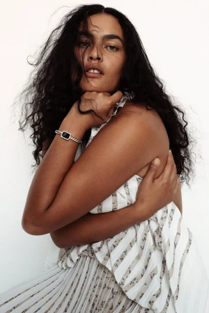 Model Ylang Messenguiral wearing a patterned pleated dress and silver bracelet against a plain white background