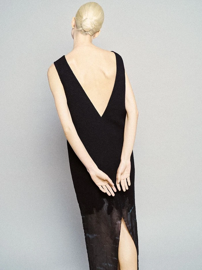 Model with blonde hair tied in a bun wearing a black V-back dress against a plain grey seamless background.