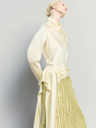 Model with blonde hair styled in a bun wearing a high neck pale yellow satin blouse and textured green skirt against a grey background