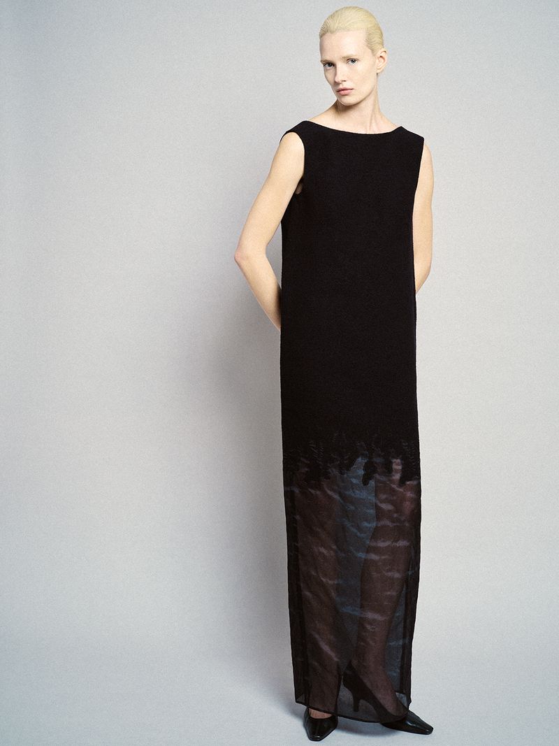 Model with blonde hair wearing a long black sleeveless dress against a grey studio backdrop