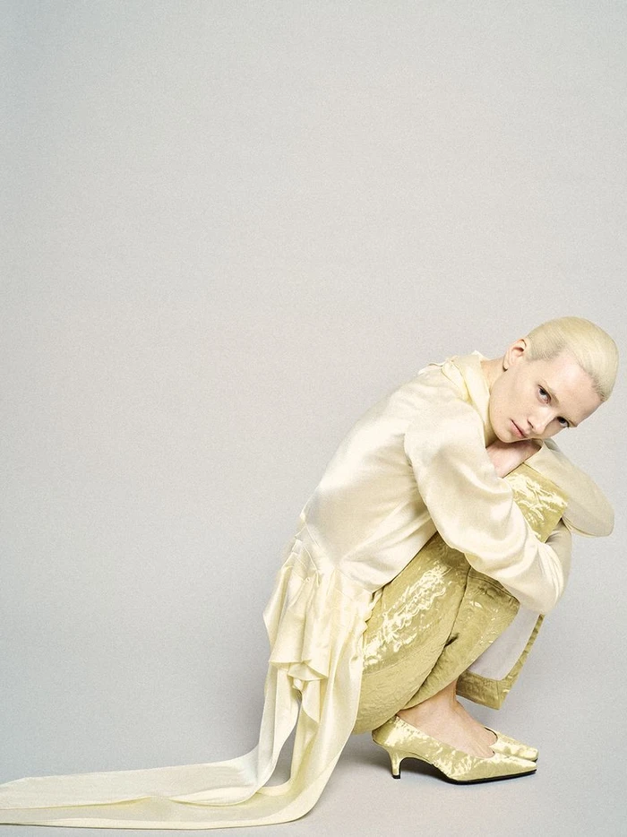 Model with blonde hair crouching against a grey background wearing a pale yellow satin blouse and textured trousers.