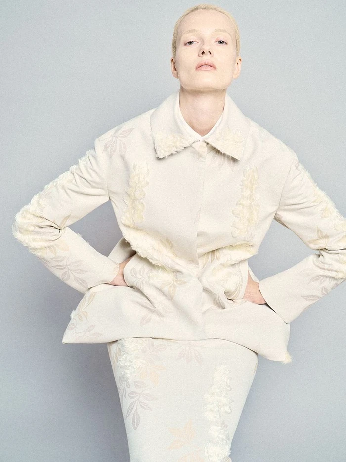 Model with short blonde hair wearing a patterned cream jacket and skirt against a grey background.