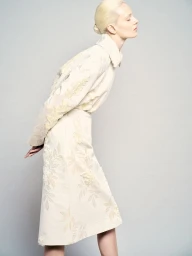 Model with blonde hair wearing a patterned cream jacket and skirt against a grey seamless background