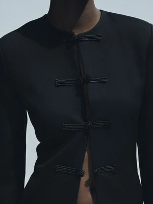 Close-up of a model wearing a black jacket with knotted cord closure details against a plain background.