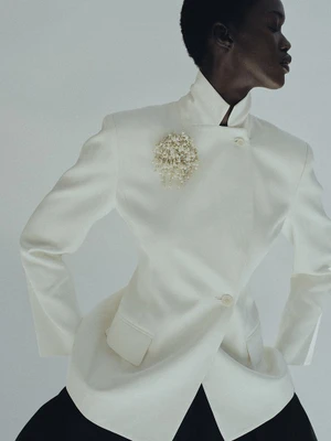 Model wearing a structured white jacket adorned with a beaded brooch against a plain studio background.