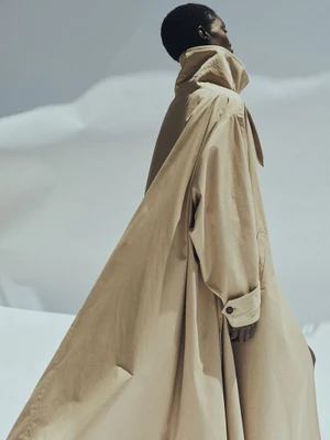 Model wearing a voluminous beige trench coat with a high collar standing against a minimalist white studio background