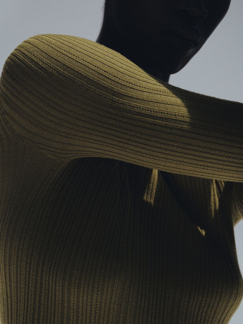 Close-up portrait of a model wearing an olive green ribbed knit top against a plain light sky background