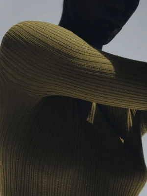 Close-up portrait of a model wearing an olive green ribbed knit top against a plain light sky background
