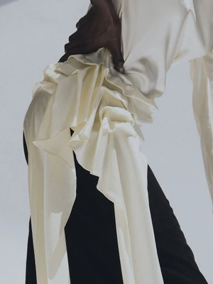 Close-up of a model wearing a draped white silk top and dark trousers against a white backdrop.