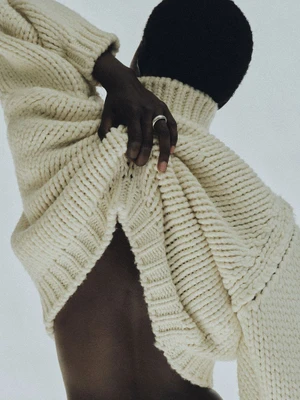 Model wearing a chunky cream knit turtleneck sweater with hand raised to the collar against a neutral background