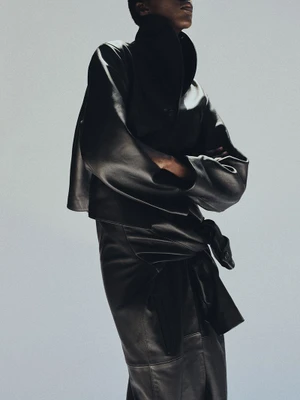 Model in a black leather jacket and matching skirt standing against a light grey studio backdrop for Kallmeyer