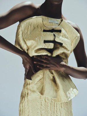Model wearing a textured yellow top with black knot closures against a pale grey background for Kallmeyer