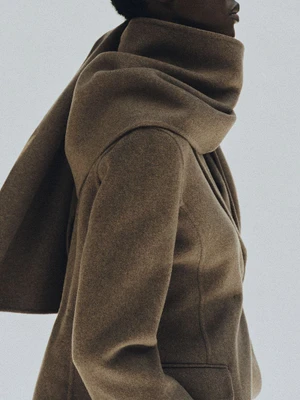 Model wearing a structured brown wool coat with an attached scarf detail against a plain grey background
