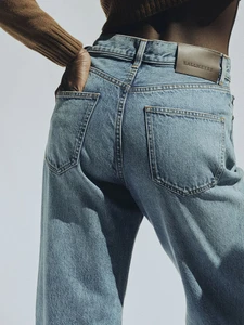 Close-up of a person wearing light blue denim jeans and a brown sweater against a neutral background for Kallmeyer