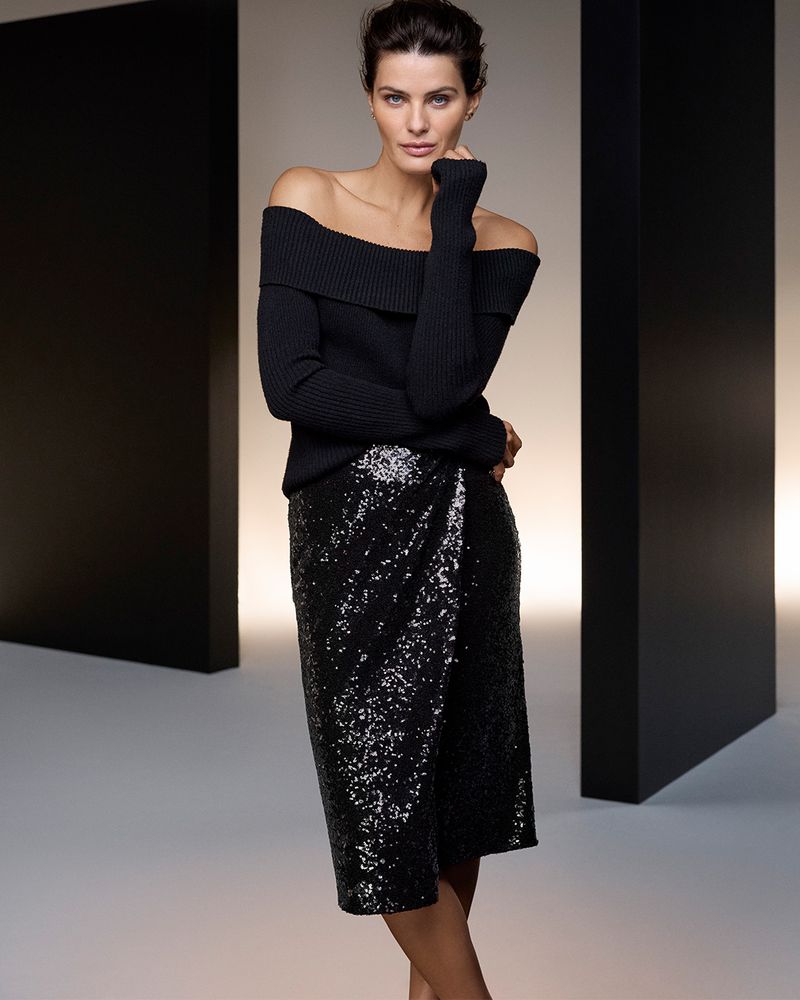 Brunette woman posing in a black off-the-shoulder sweater and sequined skirt against a studio backdrop with modern black accents.