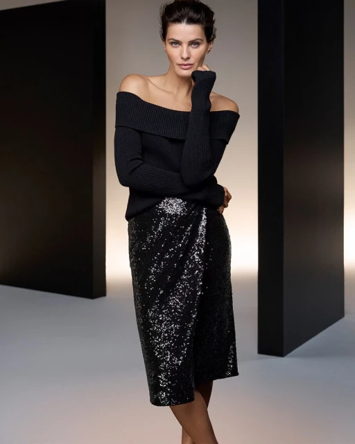 Isabeli Fontana poses in a black off-the-shoulder sweater and glittering sequin skirt beside black pillars in a studio setting.