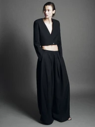 Model in a black cropped V-neck jacket and wide-leg trousers standing against a grey studio backdrop.