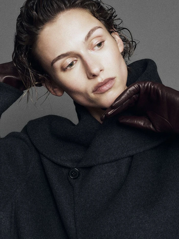 Model with slicked-back wet-look hair wearing a dark wool coat and brown leather gloves against a grey background.