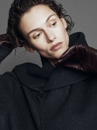 Model with slicked-back wet-look hair wearing a dark wool coat and brown leather gloves against a grey background.