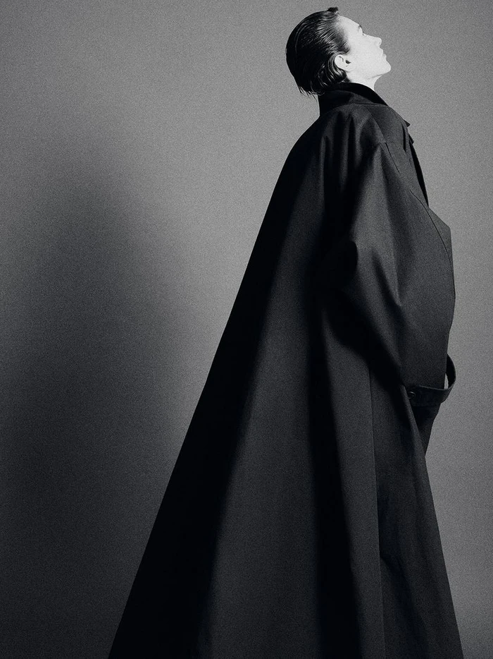 Model in a black coat with slicked-back hair posing against a neutral grey studio backdrop
