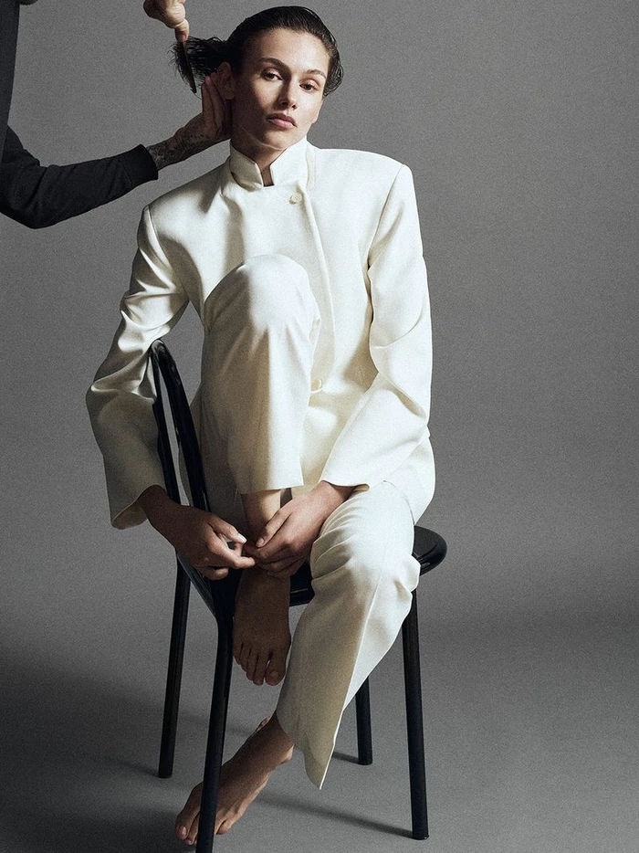 Model seated on a black chair wearing a white jacket and trousers against a grey seamless studio background.