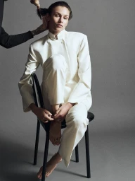 Model seated on a black chair wearing a white jacket and trousers against a grey seamless studio background.