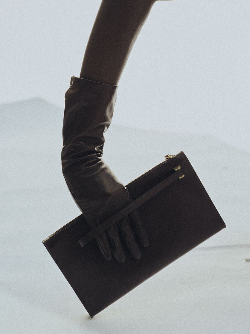 Close-up of a gloved hand holding a dark leather clutch bag against a pale background for Kallmeyer