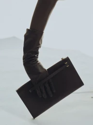 Close-up of a gloved hand holding a dark leather clutch bag against a pale background for Kallmeyer