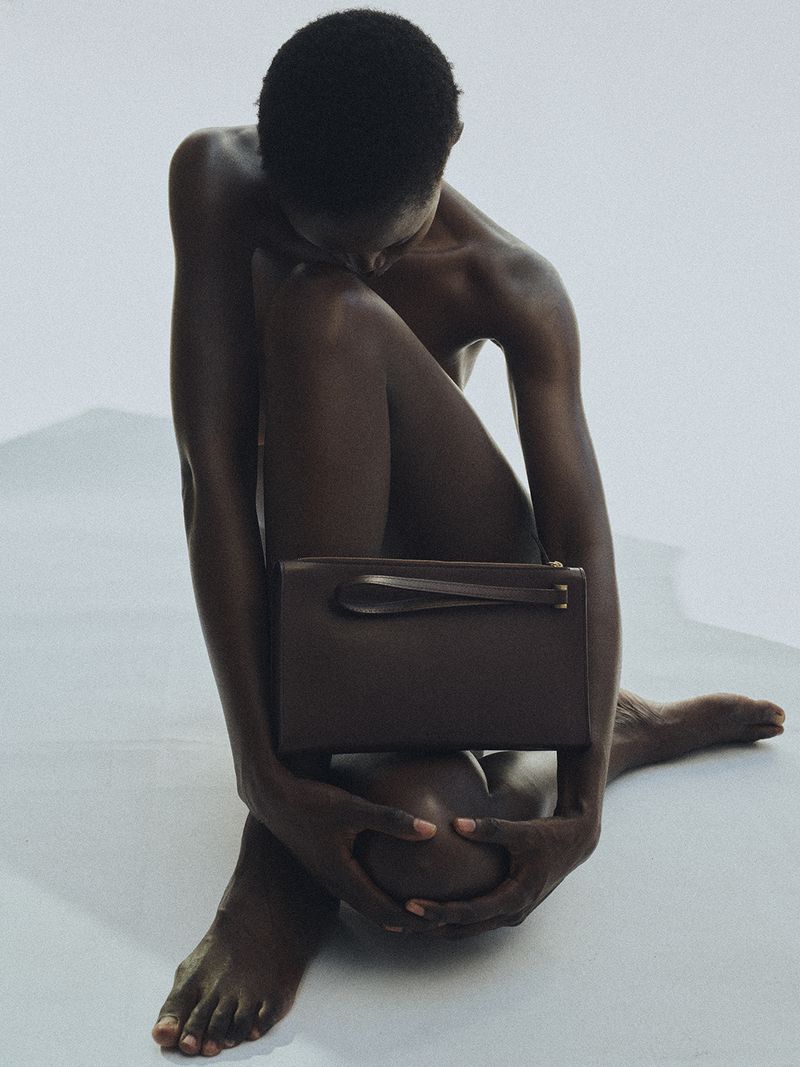 Model sitting on a white seamless backdrop holding a brown leather handbag against her knees