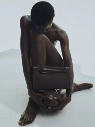 Model sitting on a white seamless backdrop holding a brown leather handbag against her knees