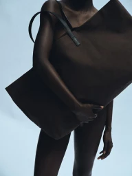 Model carrying a large dark brown canvas tote bag against a pale blue background