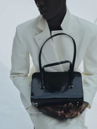 Model wearing a white structured blazer and holding a black patent leather handbag against a plain background