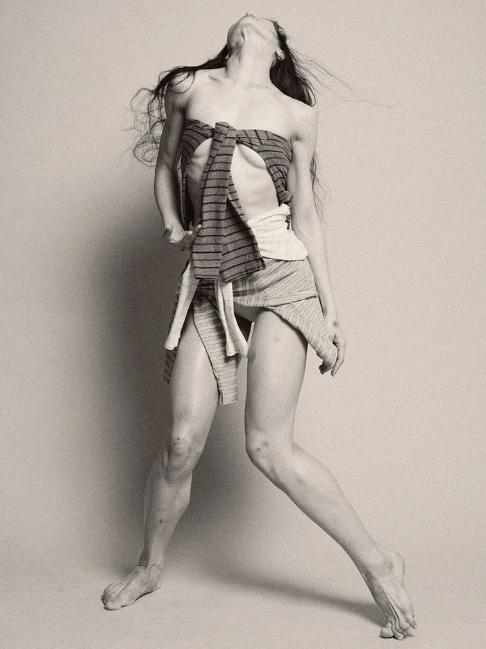 Woman in layered striped knitwear posing dynamically with head tilted back against a plain studio background.