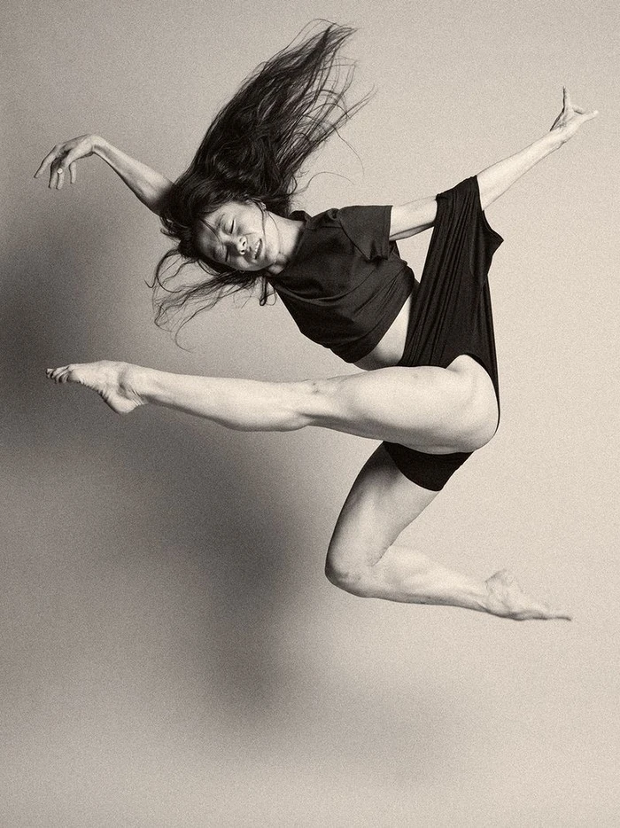 Black and white portrait of a female dancer leaping mid-air against a seamless neutral studio backdrop.