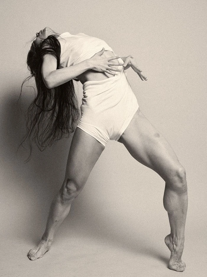 Woman arching backward in a dynamic pose, wearing a white top and shorts against a neutral studio backdrop