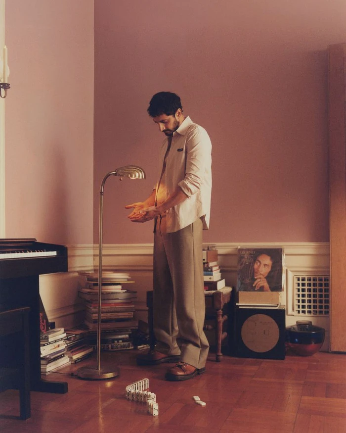 Actor Riz Ahmed standing in an interior room with a lamp and piano, wearing a light shirt and trousers.
