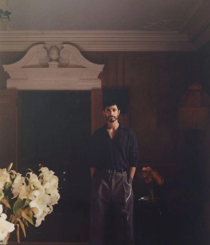 Actor Riz Ahmed standing in a dark paneled interior with white architectural molding above a doorway
