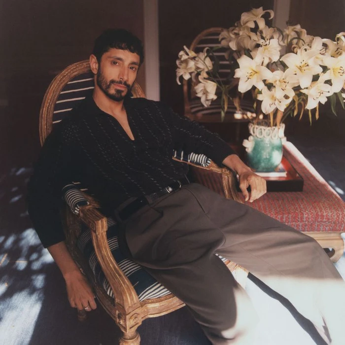 Actor Riz Ahmed seated in a patterned wooden armchair wearing a black shirt and grey trousers in a sunlit interior space